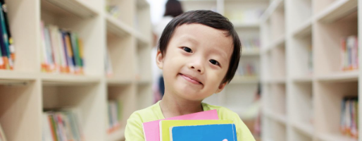 Little boy in yellow shirt in a library setting, holding three colorful books