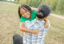 Child in green shirt hugging parent who is facing towards back in outside setting